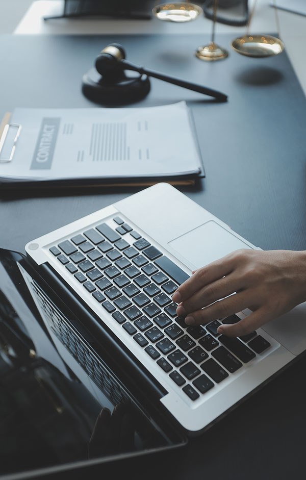 Primer plano de la mano de una persona tecleando en el teclado de un ordenador portátil, con una tableta, un mazo y documentos legales sobre un escritorio, lo que sugiere un entorno legal o judicial.